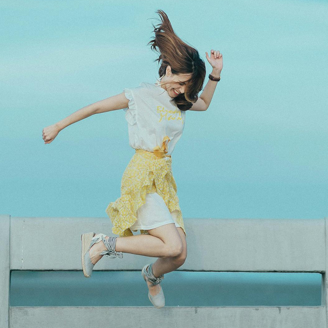 Woman in a yellow dress jumping on a wooden bridge with a blue sky background